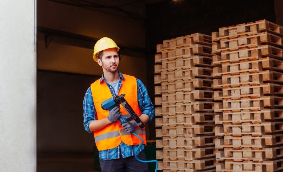 Portrait of young professional woodworker, holding pneumatic hammer, posing on stack of wooden pallets background. Indastrial tools in professional's hands. Woodworking factrory interior.
